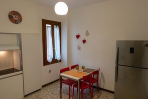 A compact kitchen with a dining table, chairs, a refrigerator, a clock, and a window with curtains.