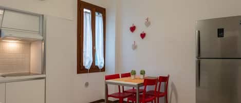 A compact kitchen with a dining table, chairs, a refrigerator, a clock, and a window with curtains.
