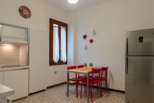 A compact kitchen with a dining table, chairs, a refrigerator, a clock, and a window with curtains.