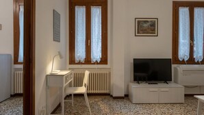 A small, well-lit room with a white desk, chair, and a television on a cabinet. There are two windows with curtains, a radiator, and a framed picture on the wall.