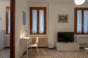 A small, well-lit room with a white desk, chair, and a television on a cabinet. There are two windows with curtains, a radiator, and a framed picture on the wall.