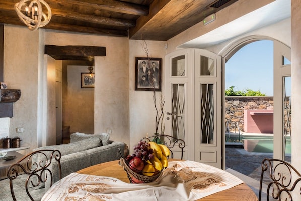 A dining area with a table set for two, a bowl of fruit, and a view of a pool through an arched doorway.
