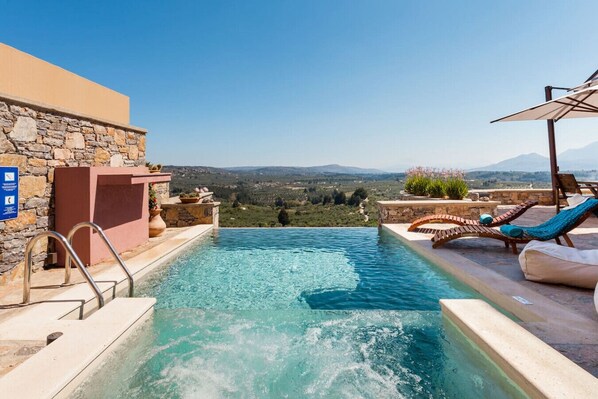 An infinity pool with a stone wall and lounge chairs, overlooking a mountainous landscape.