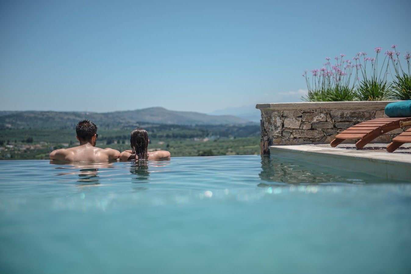 Una pareja se relaja en una piscina infinita privada con un muro de piedra y sillas de descanso, con vistas a un paisaje montañoso.