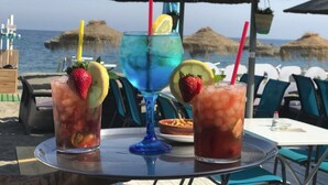 Three drinks on a table with a view of the sea and thatched umbrellas in the background. - 3-Bedroom Apartment with Sea & Mountain Views (Benahavís)