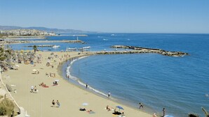 A beach with people sunbathing and swimming, a rocky breakwater, and buildings in the background. - 3-Bedroom Apartment with Sea & Mountain Views (Benahavís)