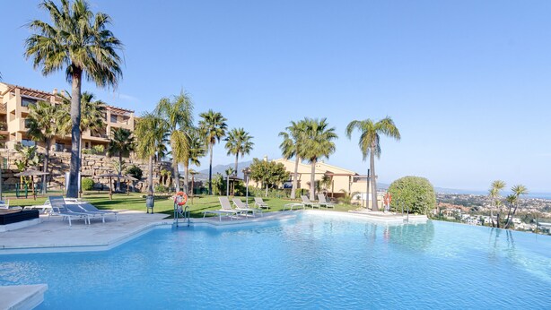 A resort pool area with palm trees, lounge chairs, and a clear view of the city in the background. - 3-Bedroom Apartment with Sea & Mountain Views (Benahavís)