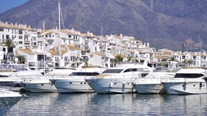 A marina with several yachts docked in front of a coastal town with white buildings and a mountain in the background. - 3-Bedroom Apartment with Sea & Mountain Views (Benahavís)