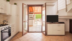 Une cuisine moderne avec des meubles blancs, une cuisinière et un four. Une porte-fenêtre coulissante donne accès à un espace extérieur aménagé avec une table et des chaises.