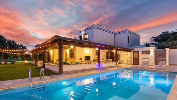 Un espace moderne aménagé au bord de la piscine, avec un coin salon abrité, une piscine et un ciel au coucher du soleil.