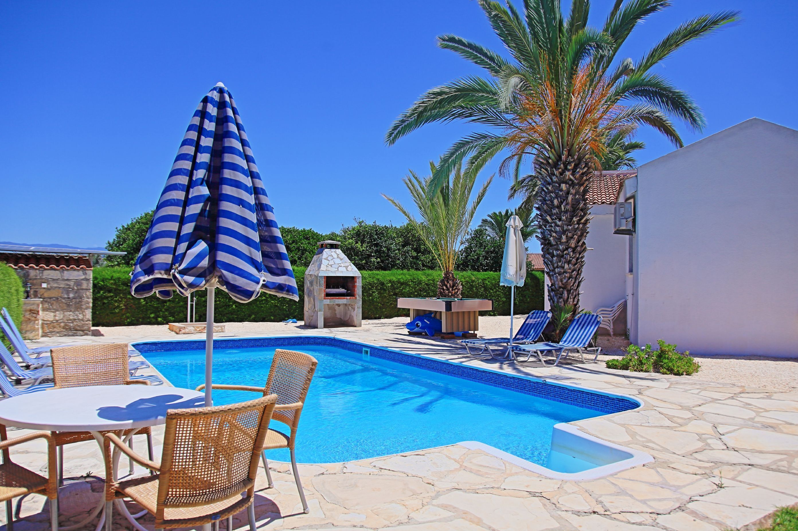 A swimming pool with lounge chairs and a table, surrounded by stone tiles and palm trees.