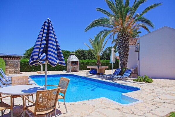 A swimming pool with lounge chairs and a table, surrounded by stone tiles and palm trees.