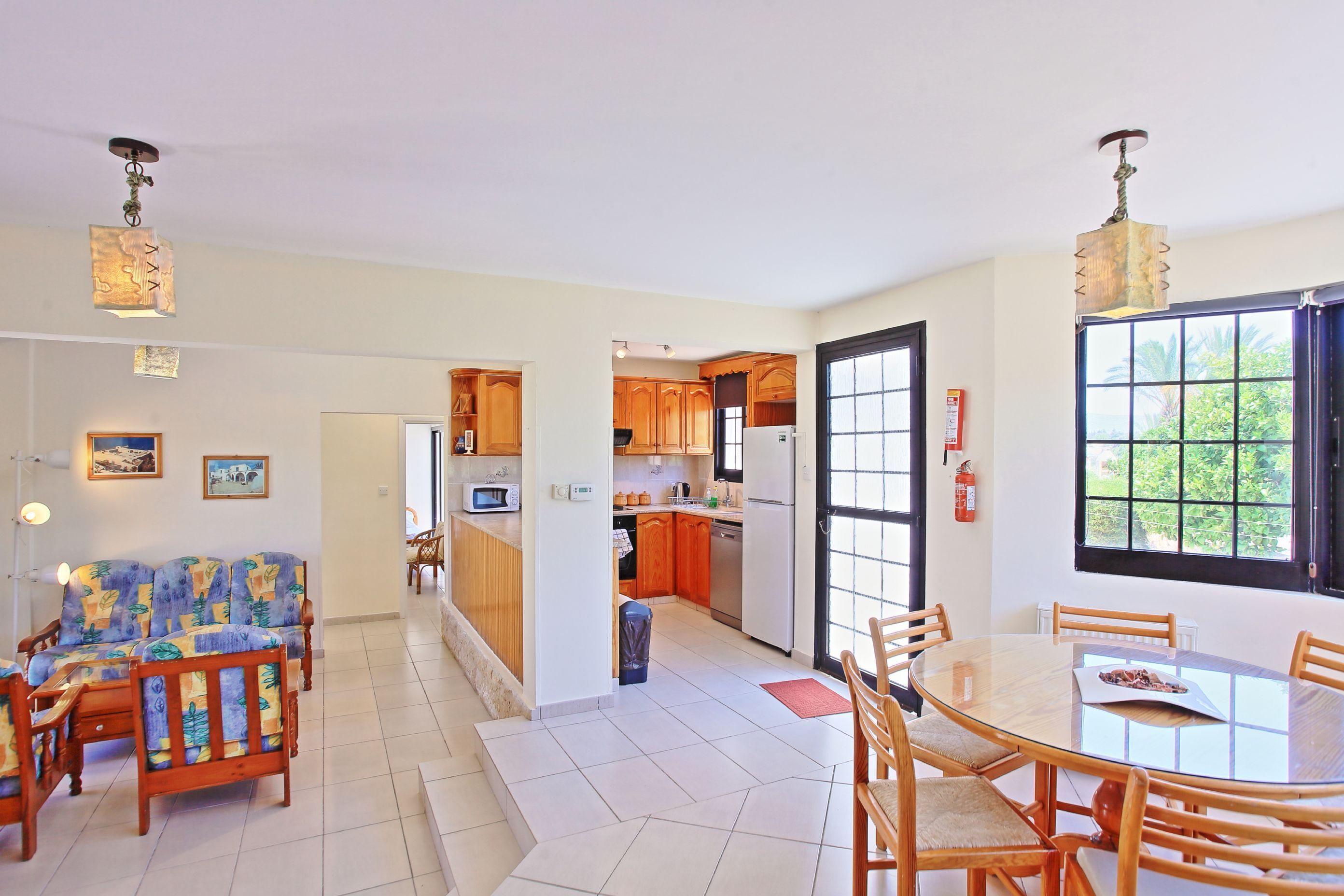 A spacious living area with a sofa and chairs, a dining table with chairs, a kitchen with wooden cabinets, and a large window with a view of greenery.