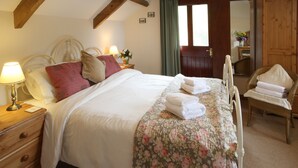 A neatly made bed with a floral bedspread and pillows, a wooden nightstand with a lamp, and a wicker chair with towels. - Courtyard cottage with extensive private grounds and free WiFi (Swansea)
