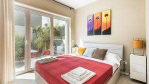 Ein modernes Schlafzimmer mit einem großen Bett, einem Nachttisch, einer Lampe und Blick auf einen Garten durch Glastüren.
