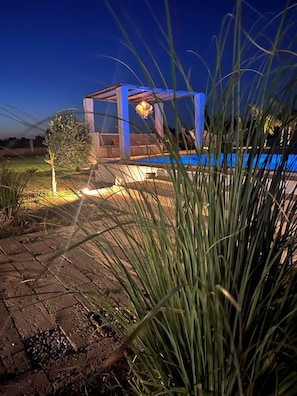 A modern outdoor pool area with a blue-lit structure, a stone pathway, and surrounding vegetation.