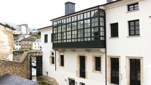 A white building with multiple windows and a large glass section. There is a stone wall and a small garden area visible.