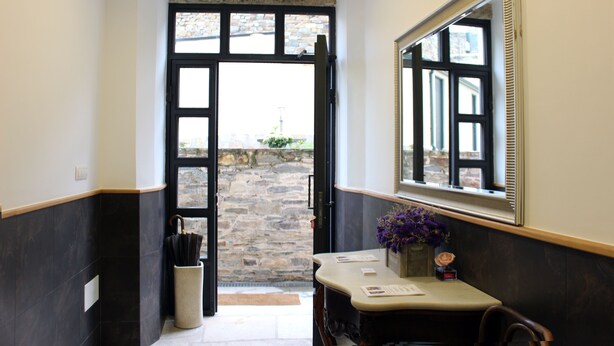 A hallway with a stone wall, a door leading outside, a mirror, and a small table with a vase of flowers.