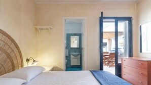 Un dormitorio con cama, cómoda, baño con lavamanos y espejo, y puerta abierta que da a un balcón.