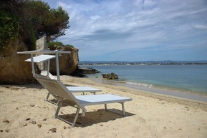 A beach with two white lounge chairs and a rocky cliff.