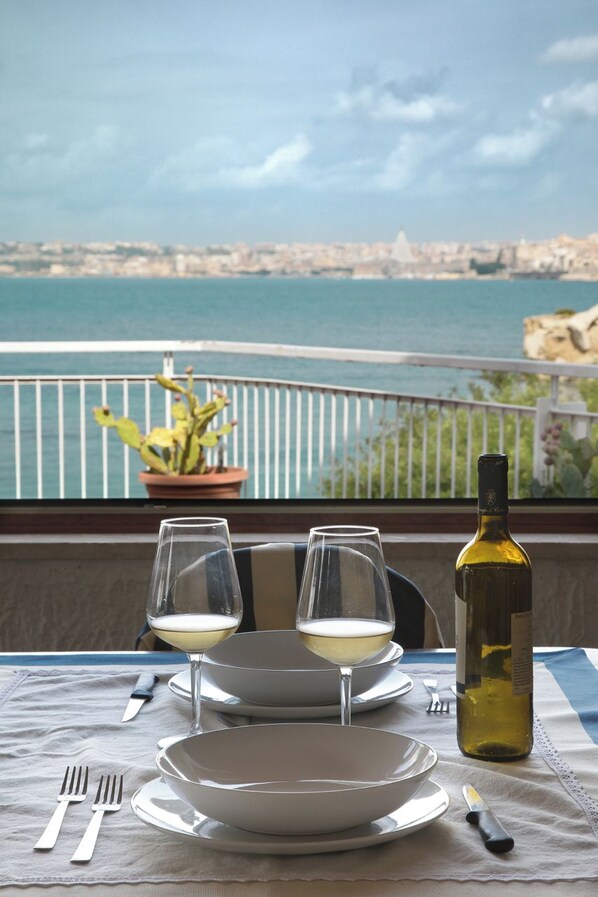 A table set with two wine glasses, a bottle of wine, and plates on a balcony overlooking a coastal cityscape.