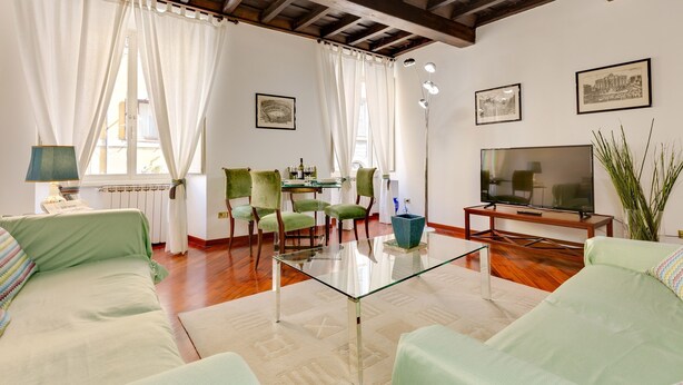A living room with a glass coffee table, green sofas, a TV, and framed pictures on the wall. - Panisperna Apartment | Comfortable and bright in Monti  (Roma)