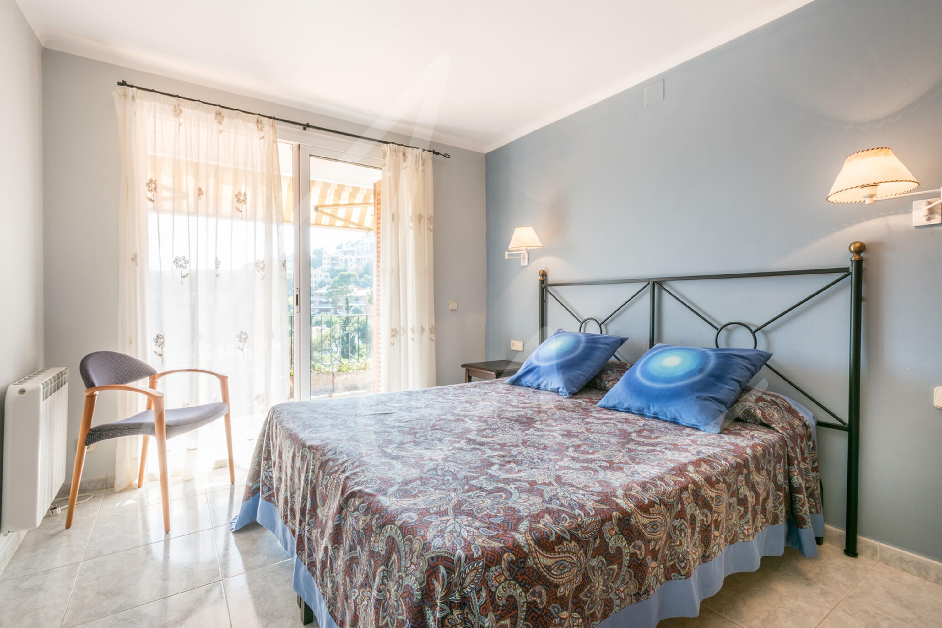A bedroom with a bed, a chair, a window with curtains, and a lamp on the wall.