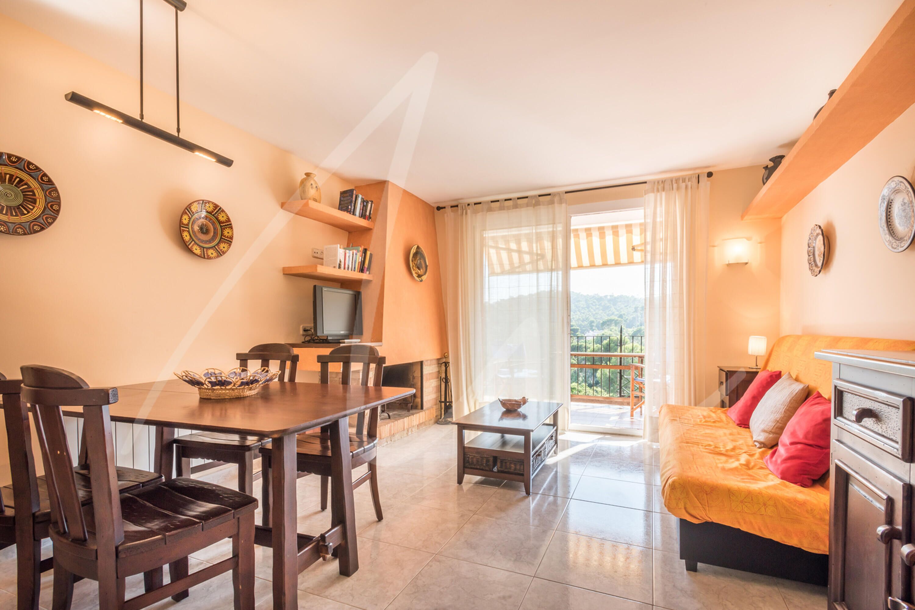 A living room with a dining area, a sofa, and a TV. There is a balcony with a view.