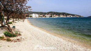 Une plage de galets bordée d’une rangée d’arbres, avec un bâtiment visible au loin.