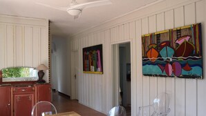 Un coin repas avec une table et des chaises en bois, un ventilateur de plafond et deux tableaux colorés accrochés au mur.