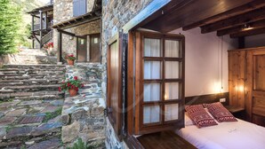 A stone building with a wooden staircase leading to a balcony. A bed with patterned pillows is visible inside.