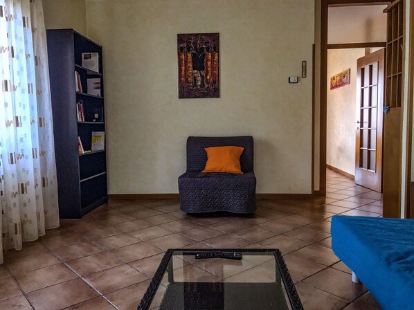 A living room with a dark blue armchair, a glass coffee table, a bookshelf, and a painting on the wall.