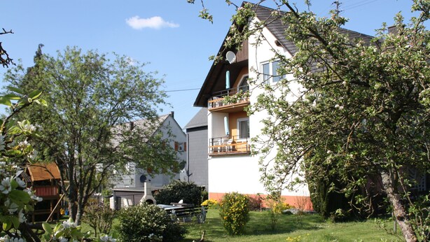 A two-story residential building with a balcony, surrounded by lush greenery and flowering trees. - Haus Buchholz Apartment 3   (Liesenich)