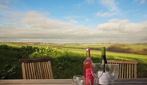 A wooden table with two wine bottles and three wine glasses on a terrace overlooking a green landscape.