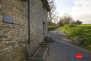 A stone cottage named "Jack's Cottage" with a gravel path leading to it, a bench, and a rustic outdoor setting.