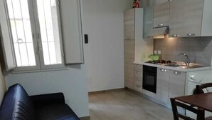 A modern living room with a dark blue sofa, a dining table with chairs, and a kitchen area with white cabinets and a stove. - Villa Barletta apartment in villa ground floor n.1 (Marina di Acate)