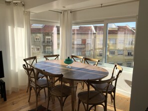 A dining area with a round wooden table and wicker chairs, large windows with curtains, and a view of neighboring buildings.