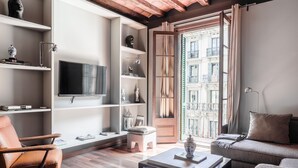 A modern living room with a flat-screen TV, wooden flooring, and a view of a building through a large window.