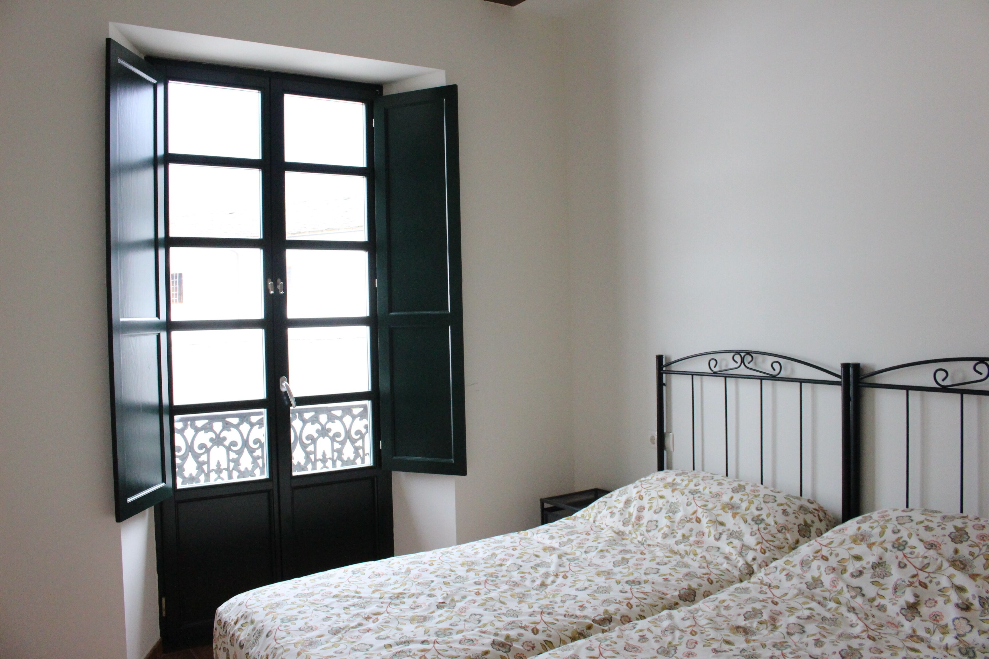 Un dormitorio con cama, una ventana con persianas abiertas y una mesita de noche.