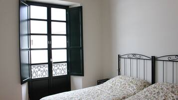 Un dormitorio con cama, una ventana con persianas abiertas y una mesita de noche.