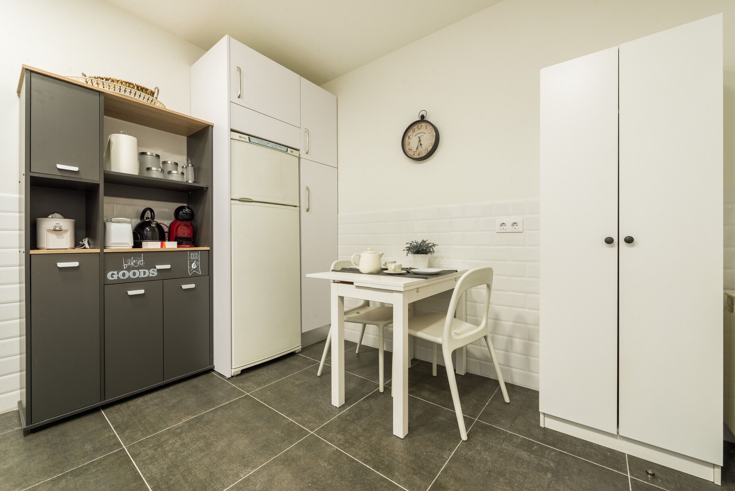 Una cocina con una mesa blanca, sillas, un reloj en la pared y un mueble con la leyenda 'productos de panadería'.