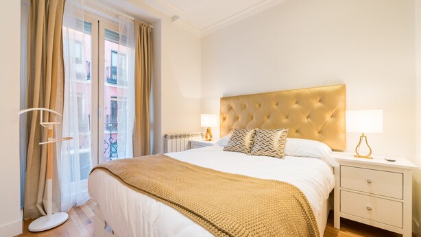 Habitación de hotel con una cama grande, una mesita de noche con lámpara y vistas al exterior a través de cortinas translúcidas.