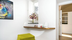 Une pièce avec un plancher en bois, un pouf vert, une étagère ornée d’un vase de fleurs, un tableau et une fenêtre donnant sur un mur de pierre.