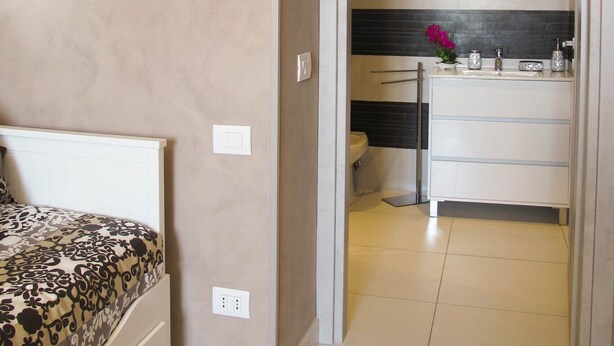 A bedroom with a bed, a bathroom with a sink and mirror, and a hallway with a framed picture on the wall.