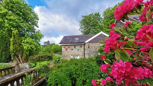 A stone building with a sloped roof, surrounded by lush greenery and a small stream.