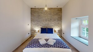 A bedroom with a stone wall, a bed with white bedding and blue pillows, a geometric-patterned blanket, and a window with a view.