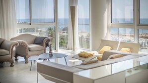 A modern living room with a sofa, armchairs, and a coffee table. Large windows offer a view of the city. - Sea view three bedroom apartment - You Stylish (Barcelona)