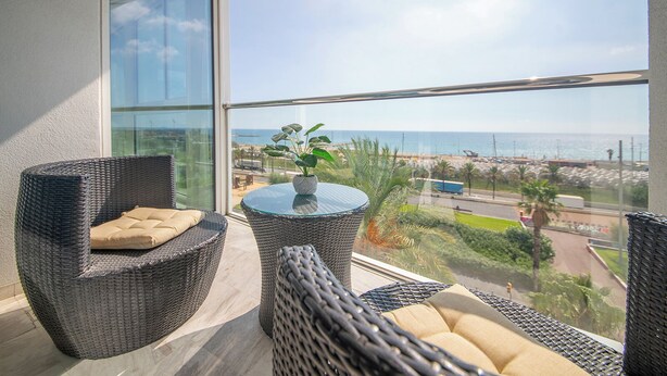 A balcony with wicker furniture and a glass table, overlooking a coastal area with a clear view of the sea and a road. - Sea view three bedroom apartment - You Stylish (Barcelona)