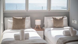 A hotel room with two beds, a bedside table with a lamp, and a view of the sea through large windows. - Sea view three bedroom apartment - You Stylish (Barcelona)