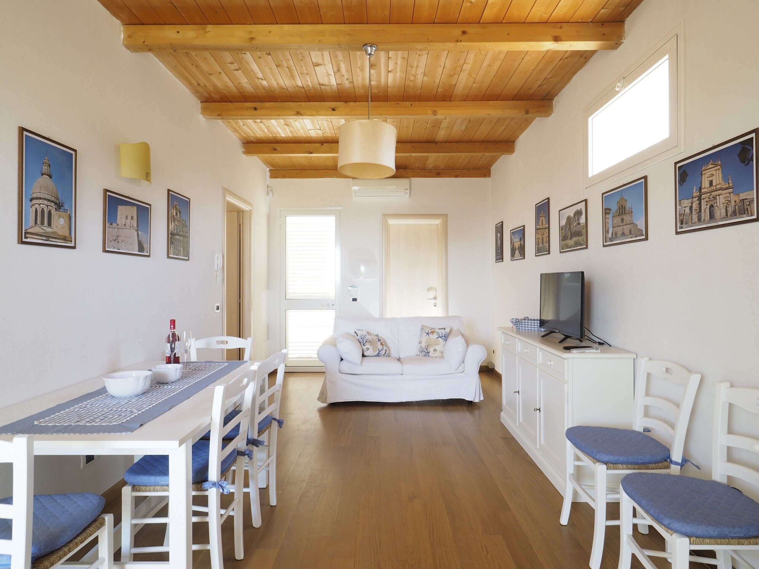 Une pièce spacieuse avec un plafond en bois, une table à manger avec des chaises, un canapé blanc, un téléviseur et des tableaux encadrés accrochés aux murs.